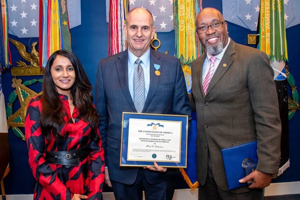 Dr. Mark Rubertone, the chief of functional information technology support for the Defense Health Agency’s Armed Forces Health Surveillance Division, receives the Department of War Distinguished Civilian Service Award during a ceremony Dec. 3, 2025, in the Pentagon’s Hall of Heroes. He was presented with his award by Dr. Merlynn Carson, left, senior advisor and chief of staff for the Office of the Under Secretary of War for Personnel and Readiness, and right, DHA Public Health deputy director Sean Friendly.