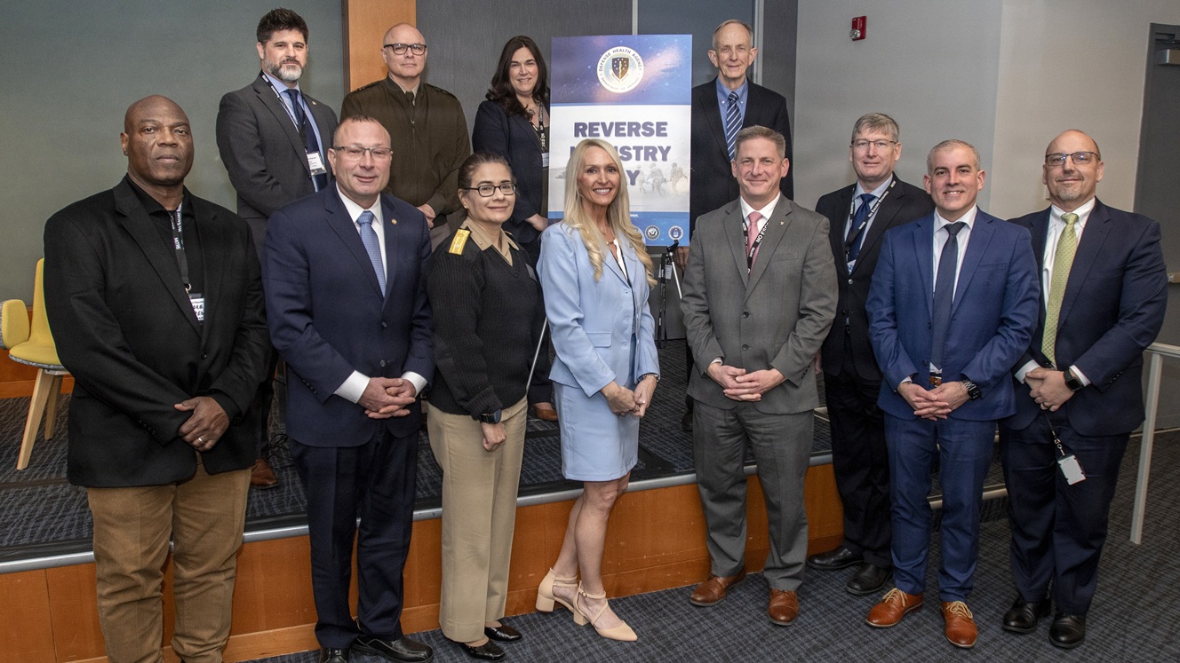 Group of people standing and posing near a stage, with a poster behind them that says "Reverse Industry Day"