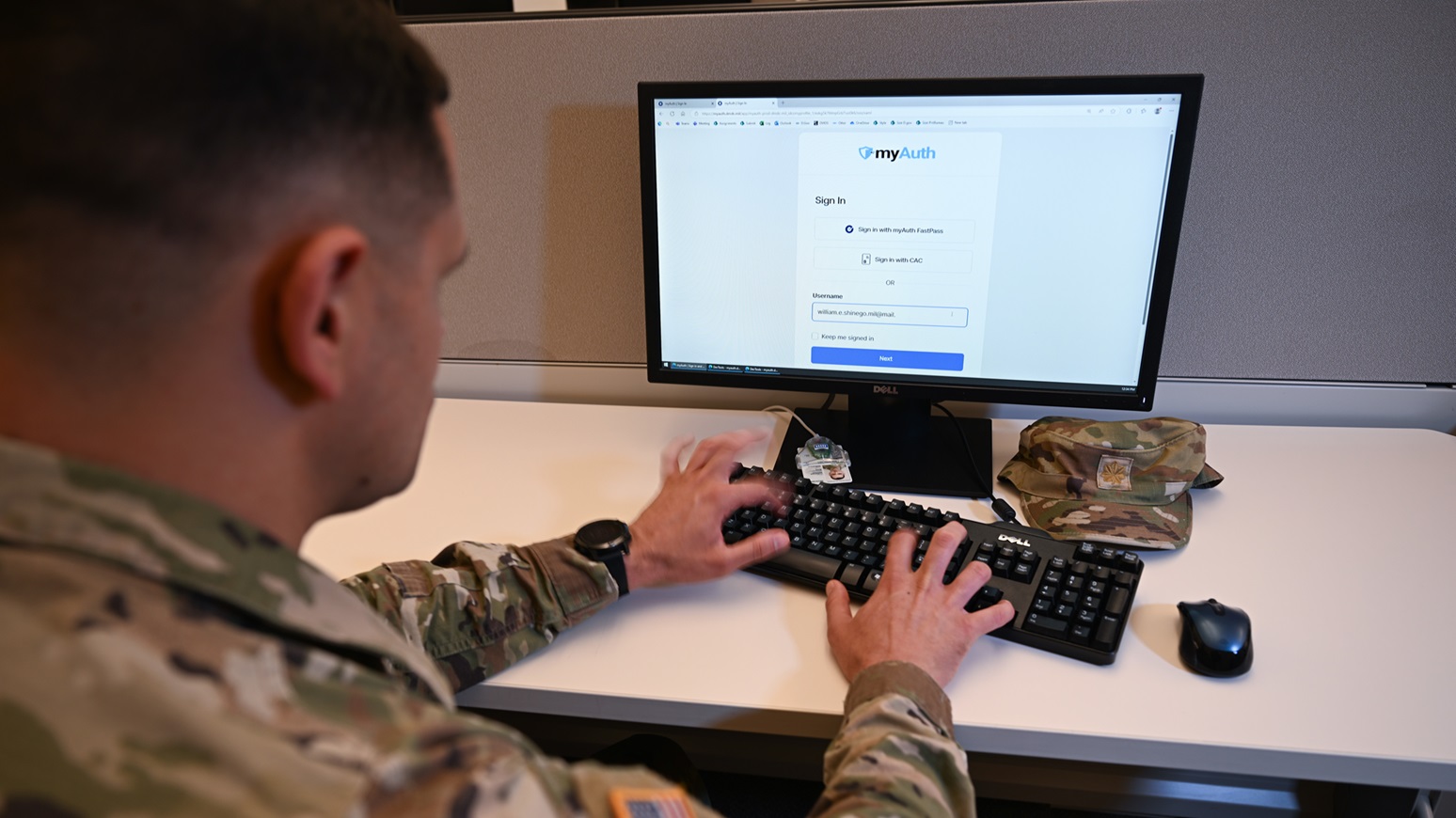 A member of the military sits at a desk and types on a keyboard attached to a monitor. 