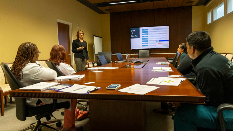 People in conference room sitting at a table while a woman makes a presentation